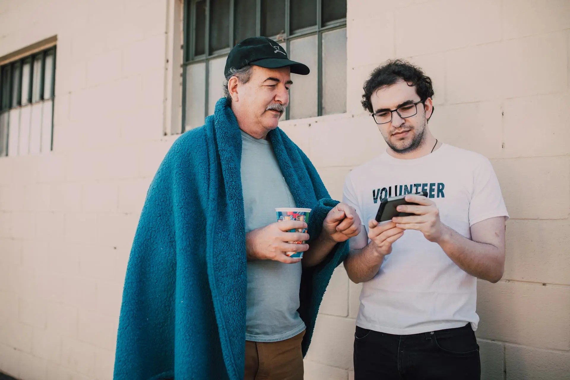 Two men stand outside near a beige wall. One, wrapped in a blue blanket, holds a cup while the other, wearing a Volunteer shirt, shares something on his phone. They seem engaged in conversation, perhaps discussing a green river story.