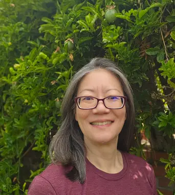 Christine Kuo, a woman with glasses and shoulder-length gray hair, smiles in front of lush green foliage. She is wearing a maroon shirt and standing outdoors.